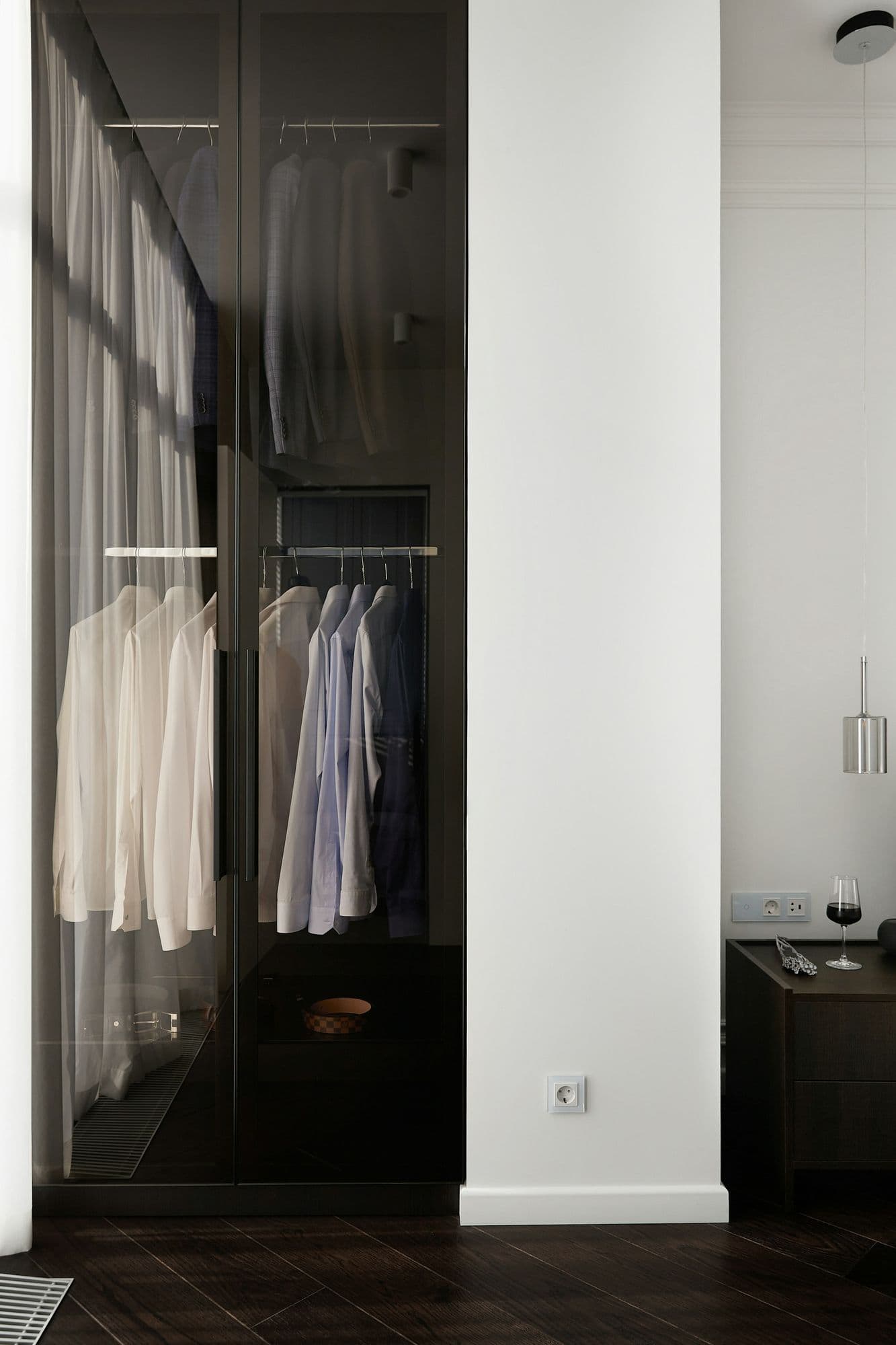 Closet com portas de vidro escuro, cabideiros iluminados e piso de madeira